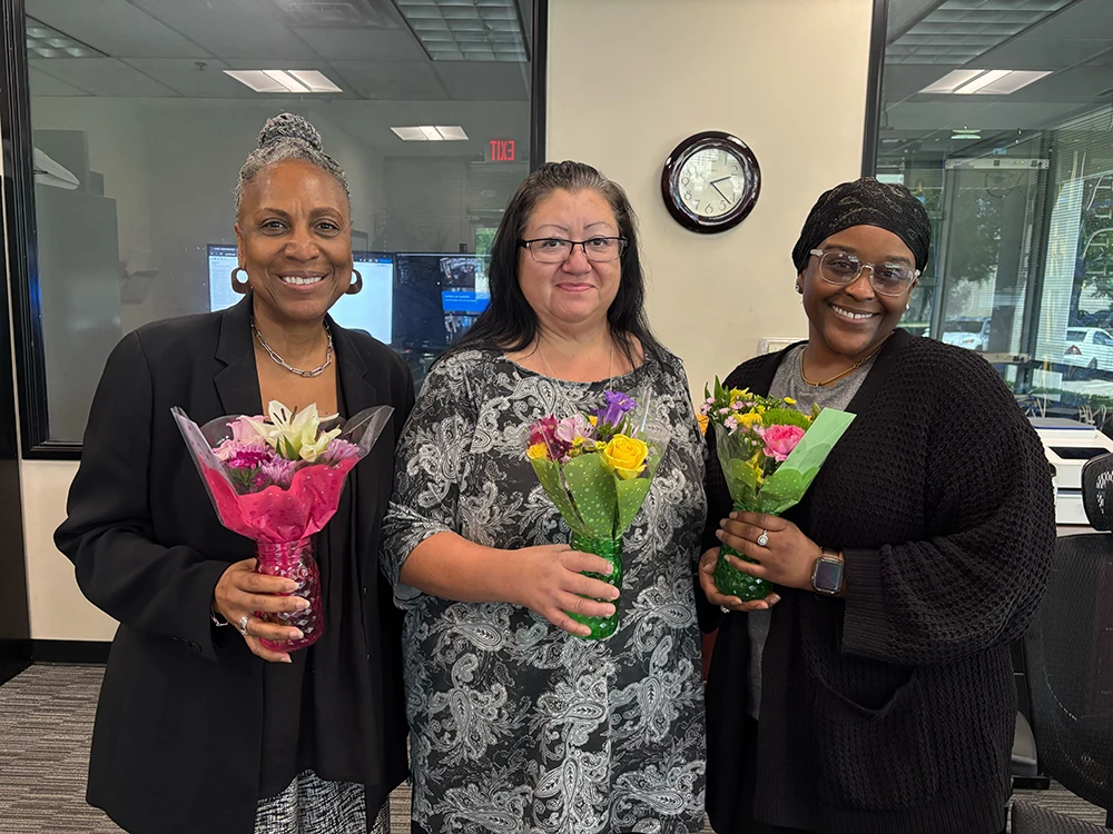 Three successful temp-to-hire placements celebrating National Staffing Week at PrideStaff Dallas - Deja (permanent receptionist), Jenny (accounting department), and Carol (accounting support) holding flowers after successful Dallas staffing agency placements