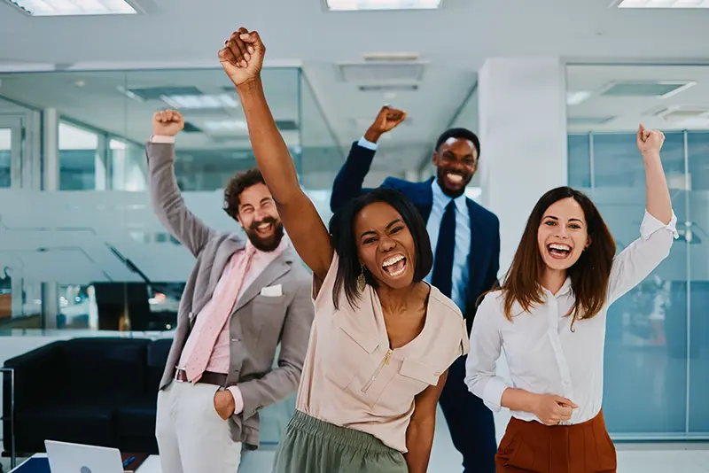 Four professionals celebrating in a modern office, arms raised in excitement — representing a successful hire with PrideStaff Dallas staffing agency.