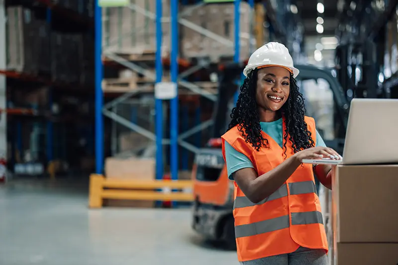 Warehouse picker in hard hat and safety vest reviewing inventory on a laptop at a Dallas distribution center