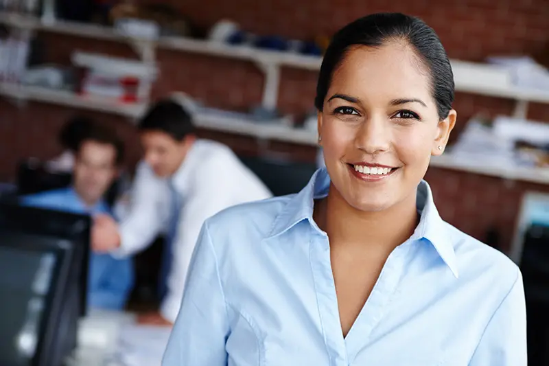 Smiling accounts payable clerk in a light blue shirt at her desk in a Dallas office, with colleagues working behind her.