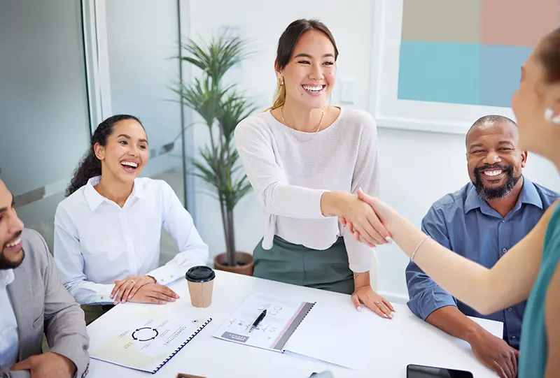 Smiling candidate and hiring manager shake hands across a conference table in a bright office.